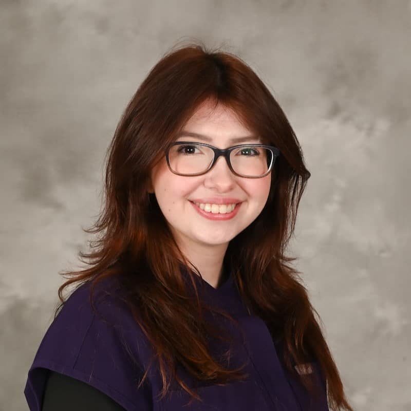 A woman with long brown hair and glasses smiles at the camera, wearing a dark purple top, against a gray studio background.