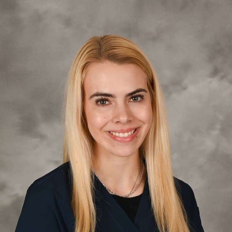 A woman with long blonde hair, wearing a dark top, is smiling in front of a gray studio background.