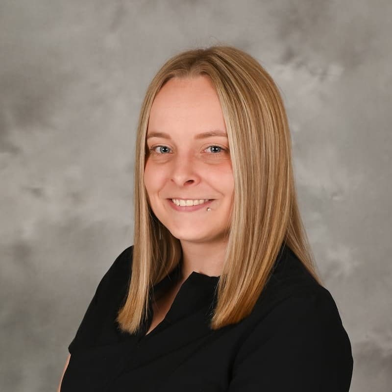 A woman with straight blonde hair wearing a black top, smiling in front of a gray mottled background.